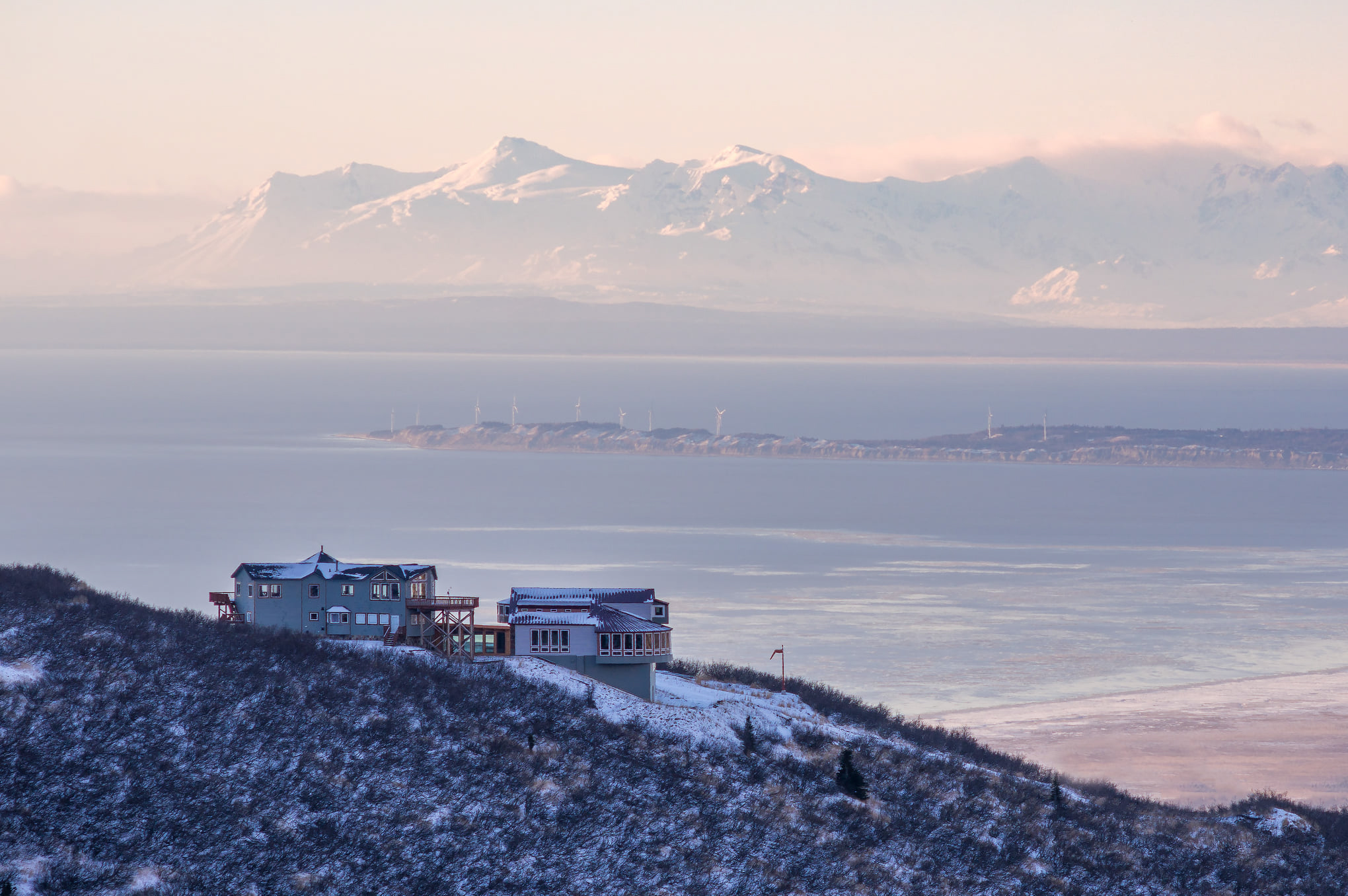 A modern home rests on a mountain ridge in winter.  The ocean and alpine mountains sit in the background.