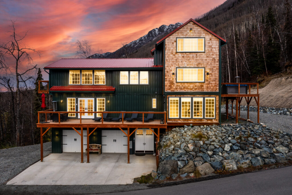 An alpine style home perched on a sloping hillside at sunset.  Warm glow emanates from the exterior windows.