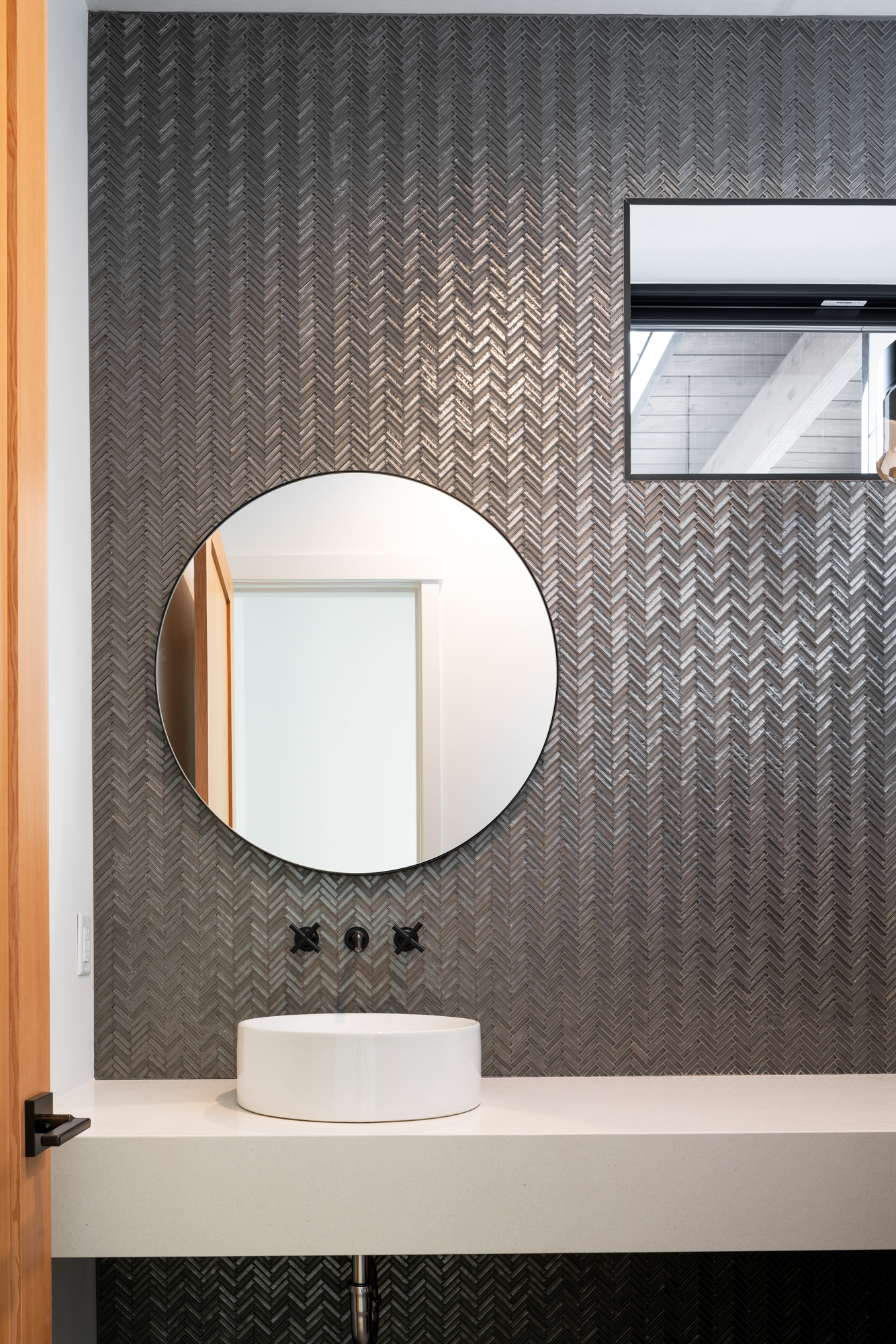 The image presents a modern bathroom vanity area, characterized by the following elements:

Mirror: A round mirror is centrally positioned against the wall, serving as a focal point.
Wall Texture: The herringbone-patterned wall provides a textured backdrop, adding visual depth.
Sink: A white, rectangular basin sink sits atop a light-colored countertop, contributing to the minimalist aesthetic.
Faucet Fixtures: Sleek, wall-mounted faucet fixtures offer a contemporary touch.
Wooden Detailing: Wooden elements to the left suggest warmth within the space.
Lighting: A small window in the upper right corner introduces natural light, enhancing the room’s textures and materials.
The composition of the photograph demonstrates a well-lit, clean, and modern space, with a focus on simplicity and elegance. The technical aspects such as lighting, focus, and composition have been executed with precision to highlight the bathroom’s design features.