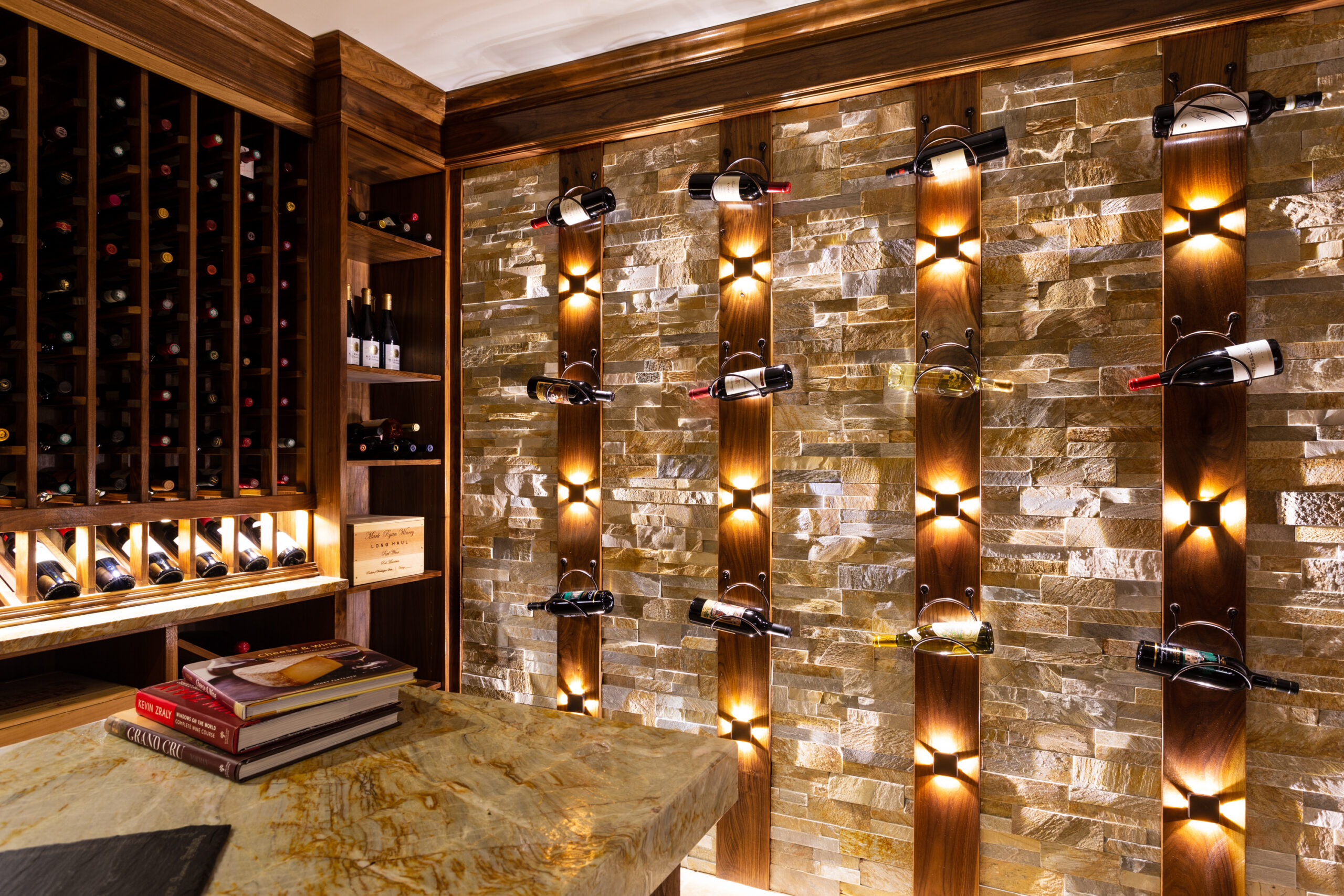 The image features an elegant wine storage area, characterized by:

Wooden Shelves: Bottles of wine are neatly displayed on wooden mounts.
Stone Wall: The backdrop of the storage area, adding a rustic touch.
Soft Lighting: Each bottle is highlighted by individual lights, creating a warm ambiance.
Marble Countertop: A sophisticated touch in the foreground with books stacked on top.
This setting conveys an upscale and inviting atmosphere for wine connoisseurs.