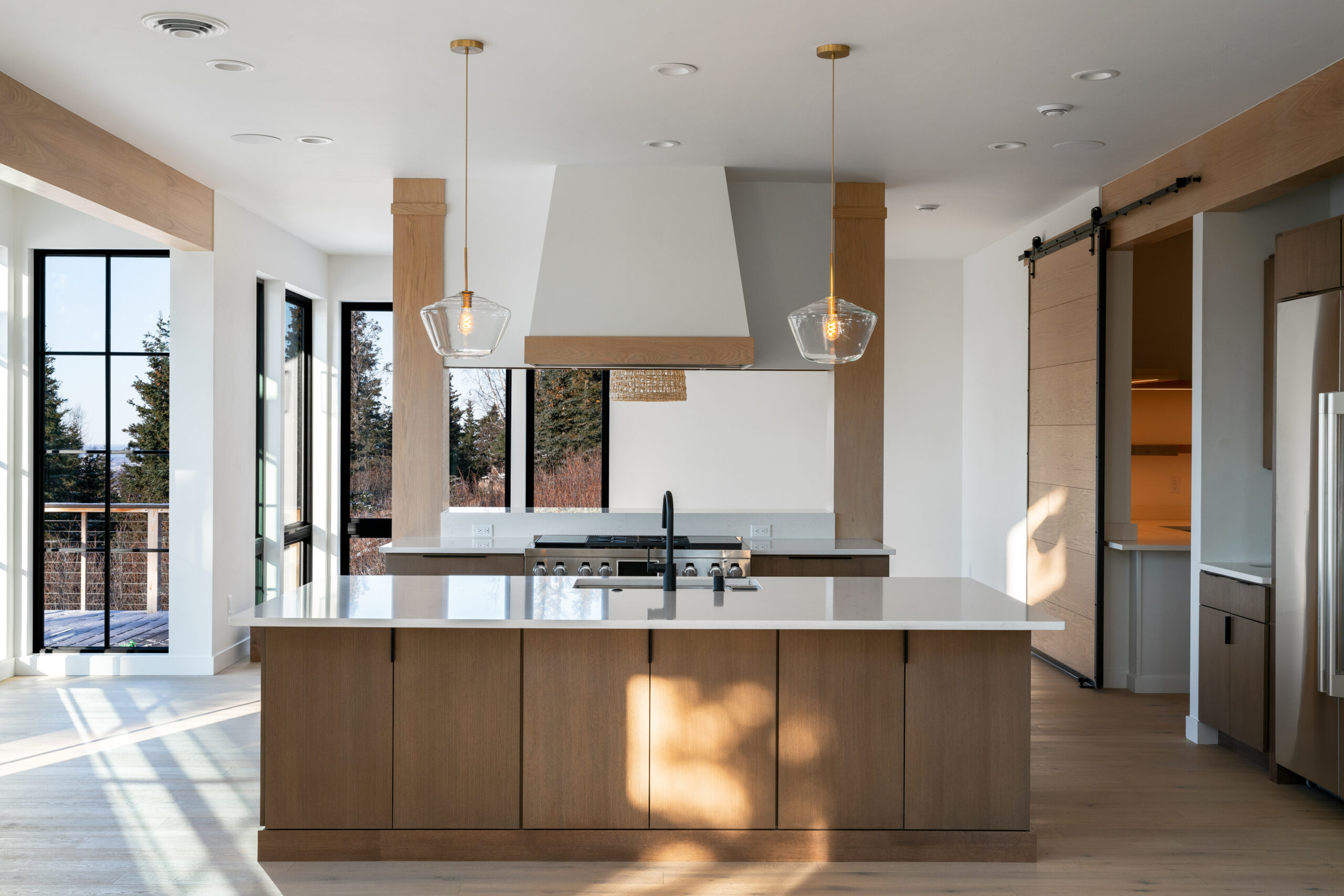 The photograph, taken by Marcus Biastock in Anchorage, Alaska, displays a modern kitchen interior with the following visual elements:

Wooden Island & White Countertop: The central wooden island is topped with a pristine white countertop, featuring a built-in sink and faucet.
Pendant Lights: Above the island, three pendant lights with gold accents provide elegant illumination.
Natural Light: Large windows allow daylight to enhance the wooden floors and cabinetry.
Stainless Steel Appliances: Integrated into the wooden cabinetry, adding a sleek touch to the space.
This image captures the essence of contemporary design, bathed in the natural beauty of Anchorage’s light. Marcus Biastock’s photography skillfully brings out the luxurious simplicity of the interior