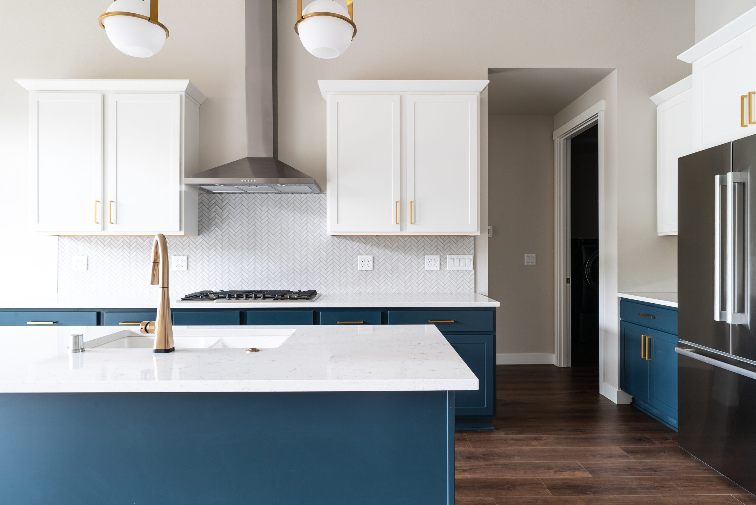 The image showcases a modern and stylish kitchen, featuring:

Two-Toned Cabinetry: Upper cabinets in white and lower cabinets including the island in navy blue.
White Countertops: Clean and polished, complementing the cabinetry.
Stainless Steel Appliances: Including a built-in stove and a large refrigerator.
Elegant Lighting: Two pendant lights with gold accents above the island.
This kitchen design blends functionality with aesthetic appeal, creating an inviting space for cooking and entertaining.