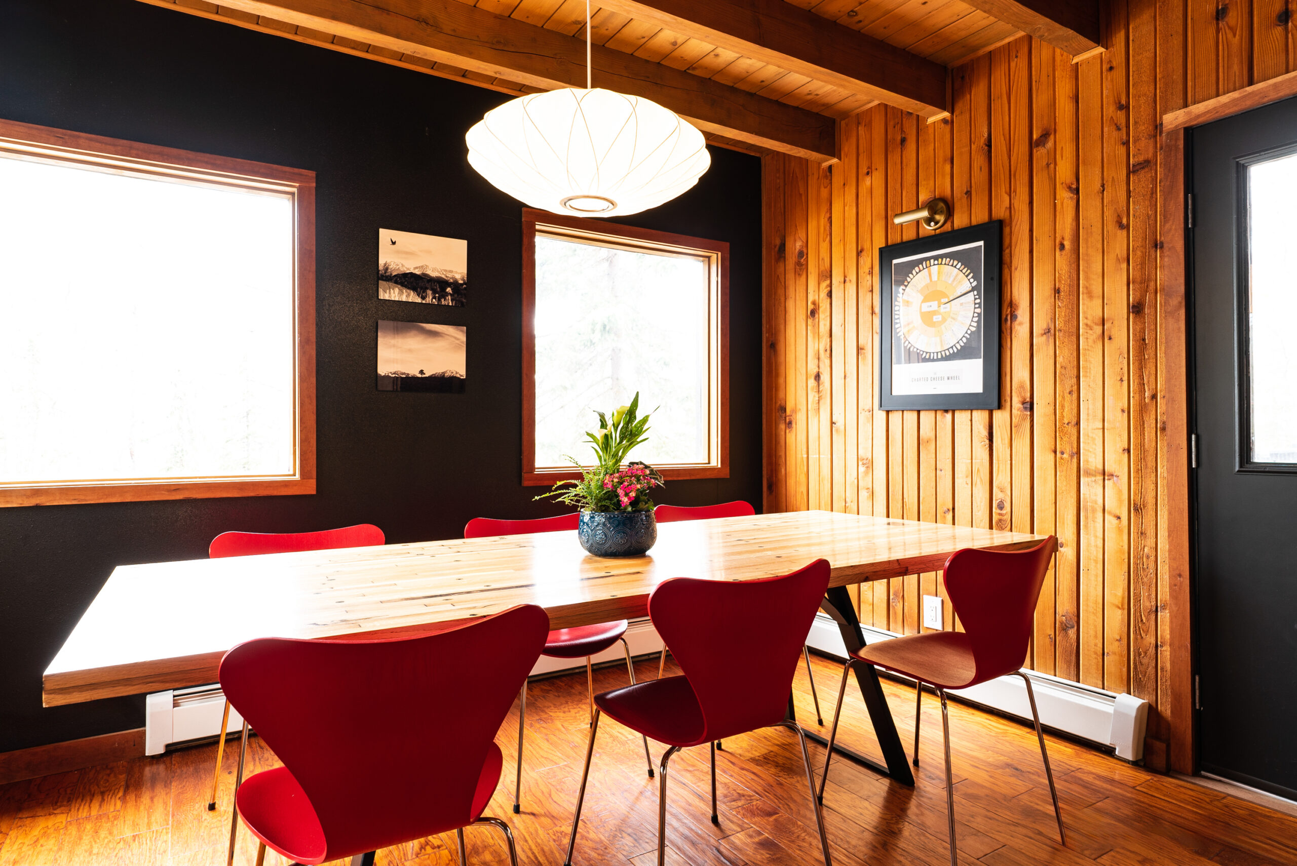 The image showcases a dining area with a modern and cozy aesthetic, featuring:

Wooden Elements: Wooden walls and a large table create a warm ambiance.
Red Chairs: Six modern design chairs add a pop of color.
Artwork: Two framed pieces adorn the walls, enhancing the decor.
Natural Light: Large windows flood the space with daylight.