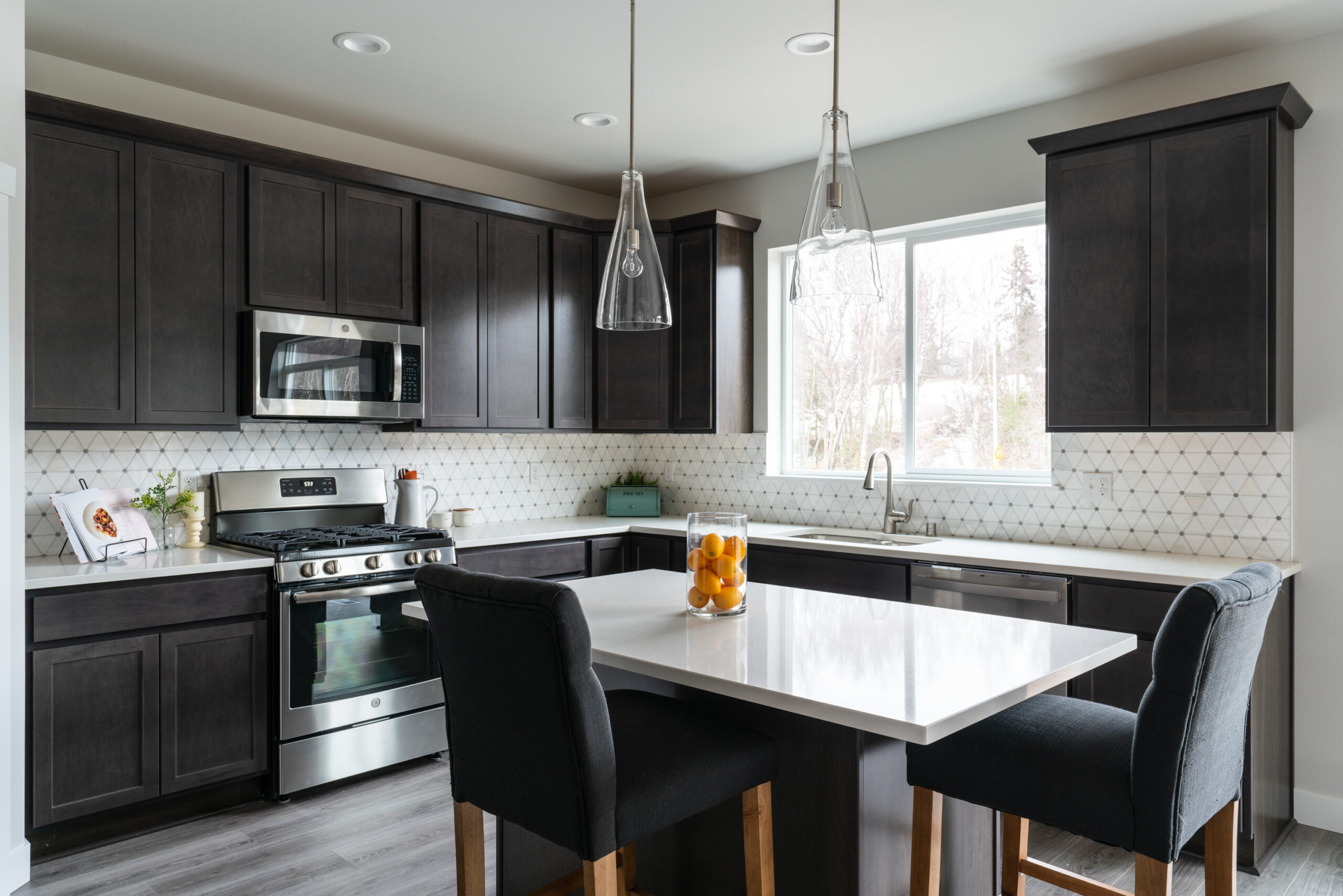 The image presents a modern kitchen that perfectly blends elegance with functionality. It features:

Dark Cabinetry: Offering ample storage and a striking contrast to the white countertops.
Patterned Backsplash: Adding texture and visual interest to the space.
Stainless Steel Appliances: Built into the cabinetry, ensuring style meets functionality.
Pendant Lights: Providing focused lighting over the island.
Natural Light: Enhancing the room’s brightness and warmth.