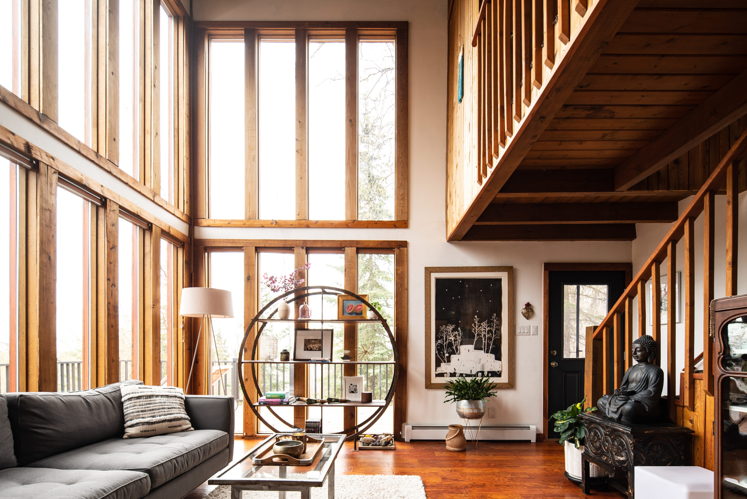 Immerse yourself in the serene ambiance of this modern living space, where natural light streams through tall wooden windows, highlighting the sleek grey sofa and unique circular shelf. Experience the perfect blend of style and comfort with our intelligent living photography.