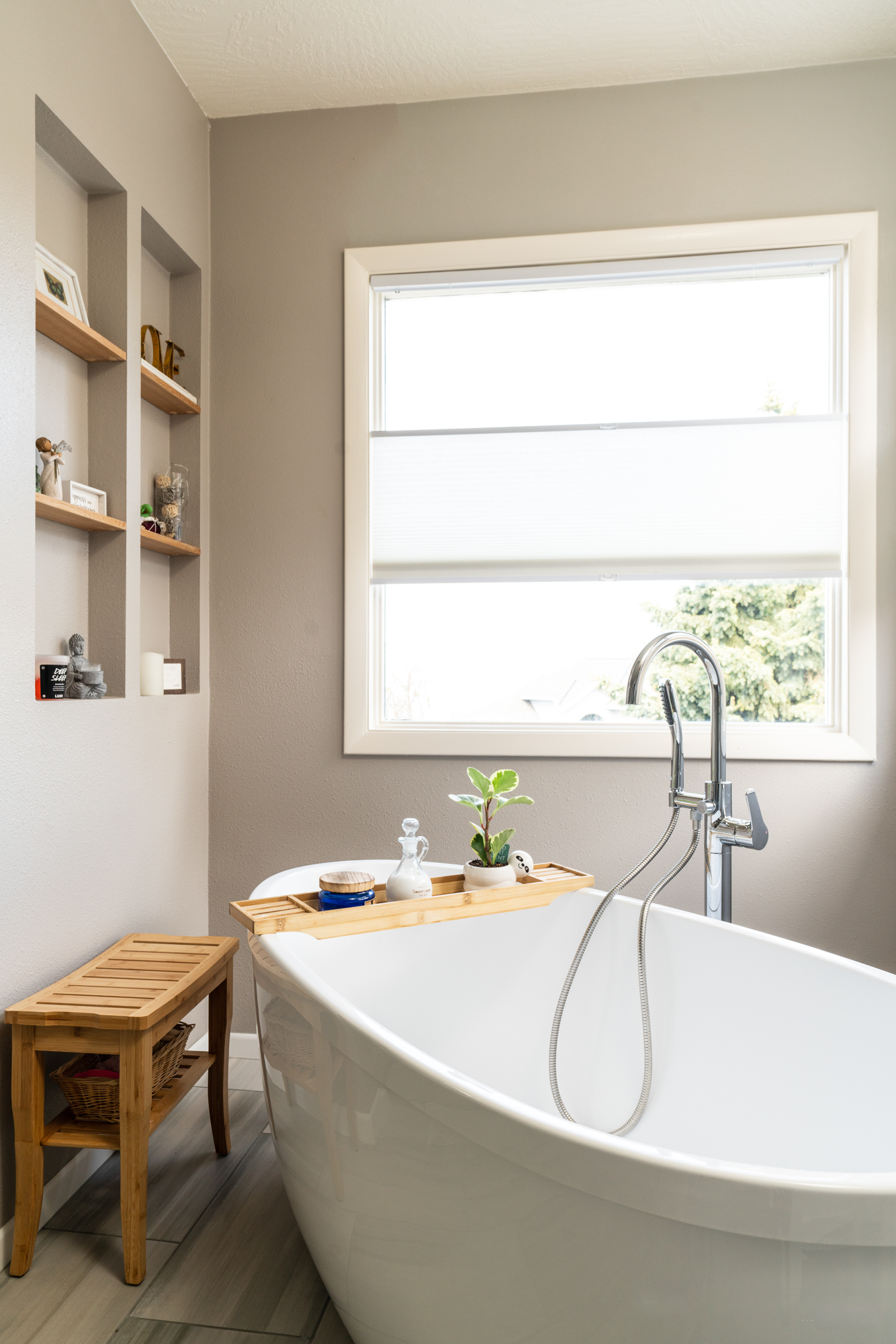 The image presents a modern bathroom setting, featuring:

Freestanding Bathtub: A sleek white tub near a large window, with a silver faucet and hand shower.
Natural Light: The window allows sunlight to brighten the space.
Wooden Stool: Positioned beside the tub, holding a blue bowl and greenery.
Built-in Shelves: Embedded in the wall, displaying bottles, jars, and decorative items.
Gray Tones: The walls and tiled floor have a light gray hue, complementing the overall aesthetic.
Photography Credit: The image was created by Photos in the House, Inc.
This description captures the essence of the bathroom’s design and ambiance.