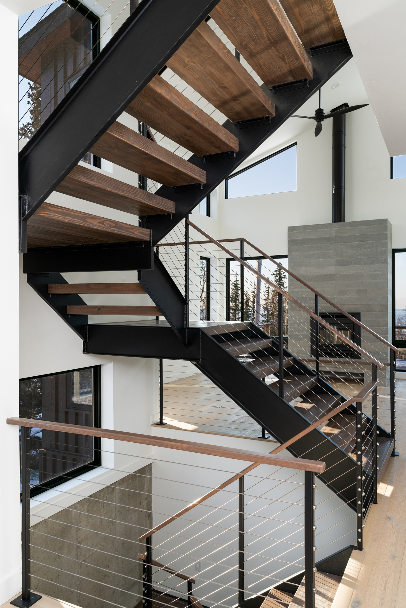 The image features a modern staircase within a spacious, well-lit area, characterized by:

Dark Metal Frames: Providing a sleek contrast to the light surroundings.
Wooden Steps: Adding a warm, natural touch to the design.
Cable Railings: Offering safety while maintaining an open feel.
Large Window: Allowing natural light to enhance the space.
White Walls: Complementing the staircase and contributing to the room’s brightness.
Ceiling Fan: Indicating a multi-level building structure.