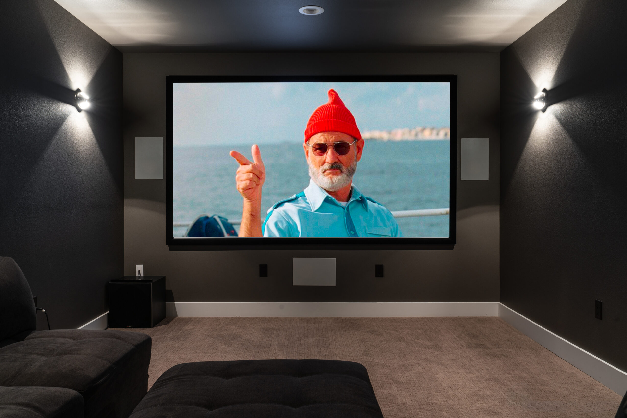The image portrays a home theater room with a distinctive ambiance, featuring:

Large Screen: Dominating one wall, displaying an image with a person making a peace sign.
Dark Walls: Creating a cinema-like atmosphere, enhanced by modern lighting.
Comfortable Seating: Plush black armchairs offer a prime viewing experience.
Additional Details: A subwoofer and wall-mounted lights are visible, completing the home theater setup.
This description provides a glimpse into the room’s design and functionality, perfect for a cozy movie night
