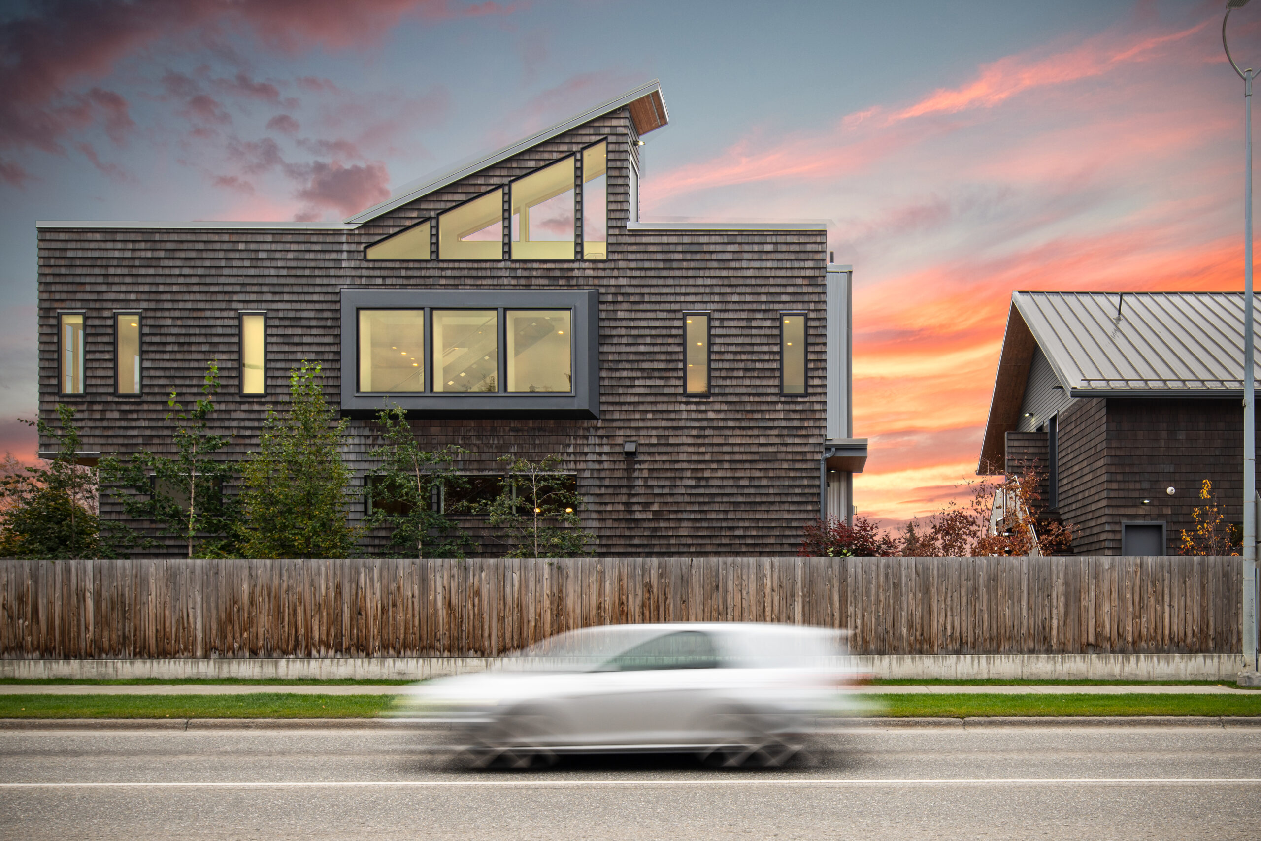 A modern house with wood shingle siding and geometric windows stands out against a vibrant sunset sky in Seattle. A car blurs by on the street in the foreground, adding a sense of motion.