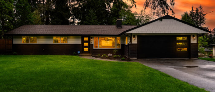 Twilight photo of a single-story house with interior lights on, creating a warm and inviting atmosphere. The sky is painted with hues of orange and pink, suggesting the photo was taken during sunset or sunrise. The house features a dark roof, light-colored siding, and an open garage. A well-maintained lawn with a visible sprinkler on the left side is in front of the house.” This alt text provides a detailed description of the image for those who might not be able to see it. It captures the key elements of the photo, including the time of day, the house’s appearance, and the surrounding environment.