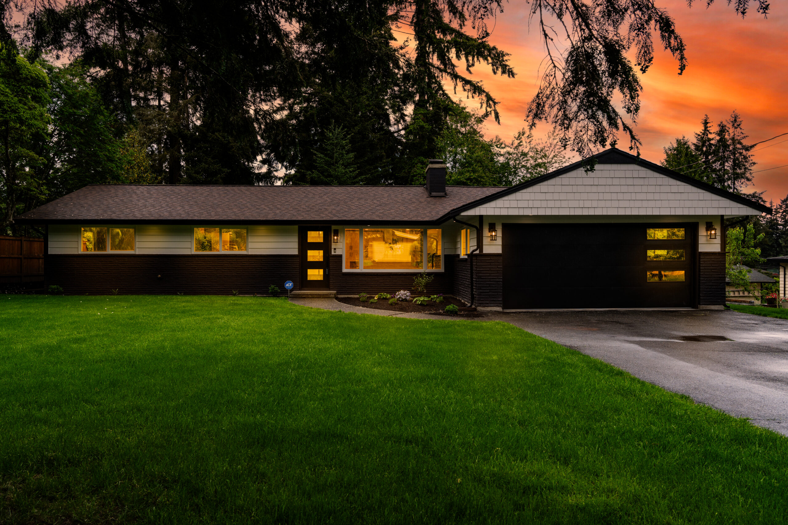 Twilight photo of a single-story house with interior lights on, creating a warm and inviting atmosphere. The sky is painted with hues of orange and pink, suggesting the photo was taken during sunset or sunrise. The house features a dark roof, light-colored siding, and an open garage. A well-maintained lawn with a visible sprinkler on the left side is in front of the house.” This alt text provides a detailed description of the image for those who might not be able to see it. It captures the key elements of the photo, including the time of day, the house’s appearance, and the surrounding environment.