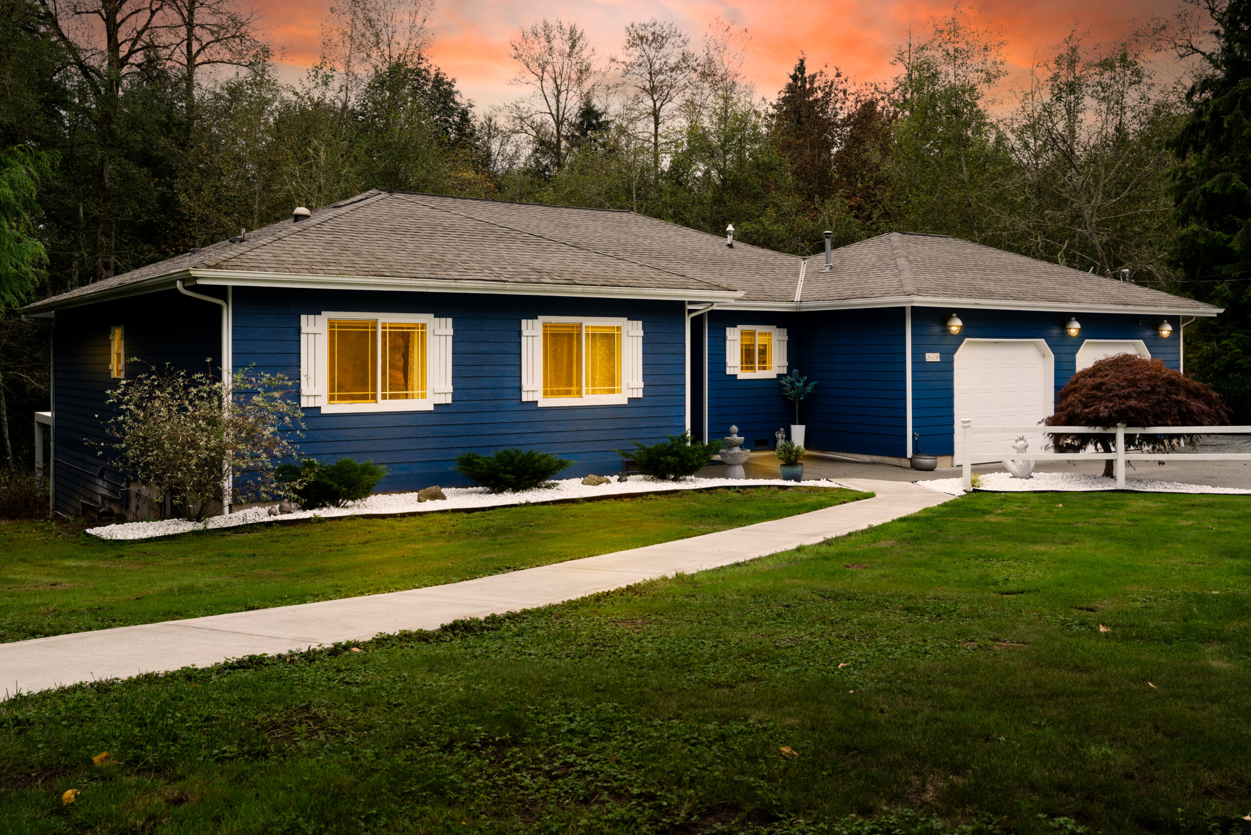 A charming single-story house with a blue exterior and white trim. The house features three windows with yellow shutters and a white front door. A well-kept lawn and a curved pathway lead to the porch. A large, rounded bush is on the right side of the house, while a small tree or shrub is on the left. The sky above is painted with hues of orange and pink, indicating either sunrise or sunset.