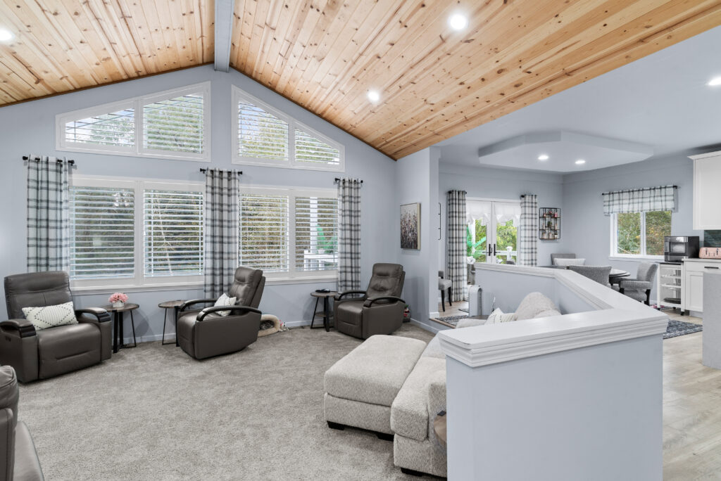 Interior of a modern living room with vaulted wooden ceilings, multiple windows with partially closed blinds, and natural light filtering in. The room features two dark leather armchairs, a gray fabric sofa, a matching ottoman, and a small glass-top coffee table. A white kitchen counter is visible in the background, and the walls are painted light blue, complementing the neutral color palette of the furniture and carpeted flooring.
