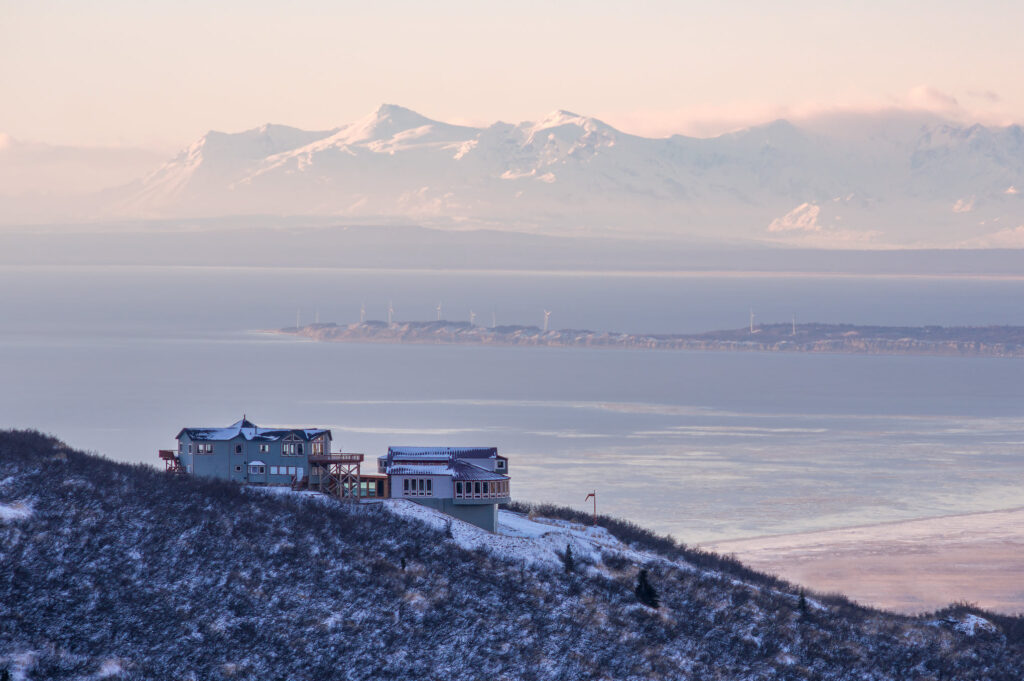 A serene winter scene featuring two modern houses perched on a snow-dusted hillside. The houses, with their distinct designs, overlook a vast, calm body of water that stretches towards the horizon. In the distance, a majestic range of snow-capped mountains rises against a soft, pastel sky, adding a dramatic backdrop to the tranquil setting. The overall atmosphere is peaceful and remote, highlighting the natural beauty and isolation of the location.