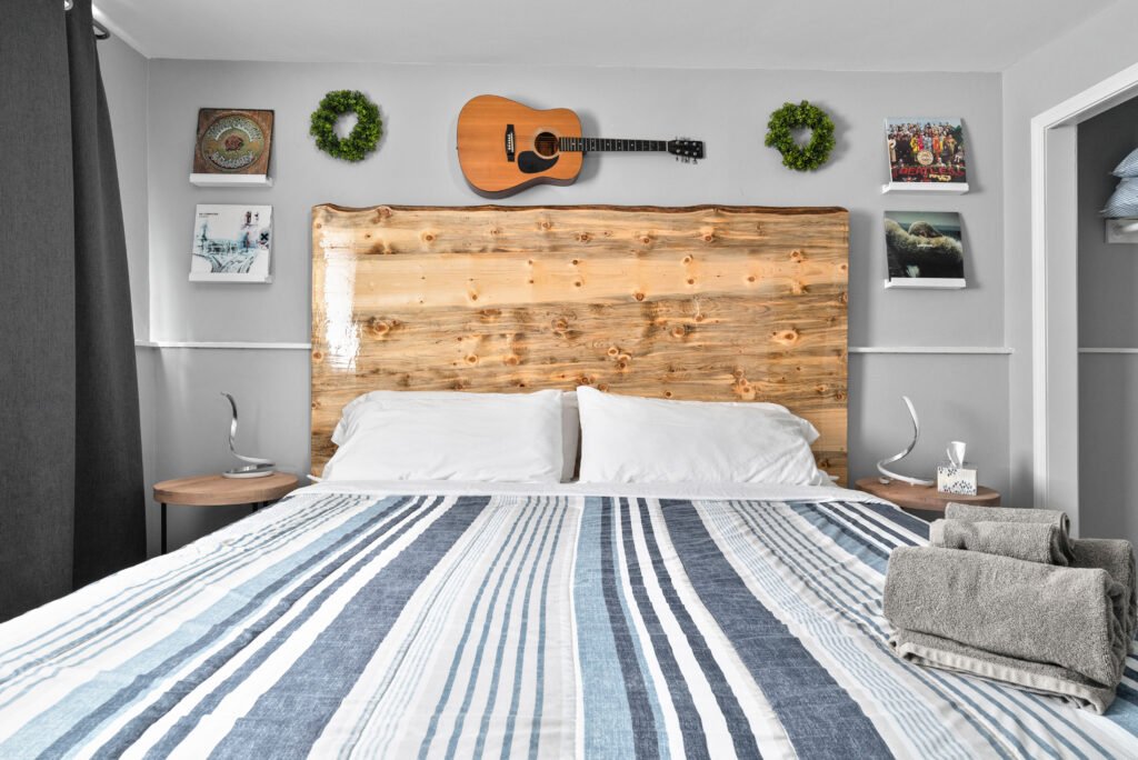 A cozy bedroom setup with a rustic wooden headboard. Above the headboard, a guitar is mounted on the wall, flanked by two green wreaths. On either side of the headboard, there are shelves with framed artwork and posters. The bed is neatly made with white pillows and a blue and white striped comforter. On each bedside table, there are modern lamps and a box of tissues on the right. At the foot of the bed, there are folded grey towels.