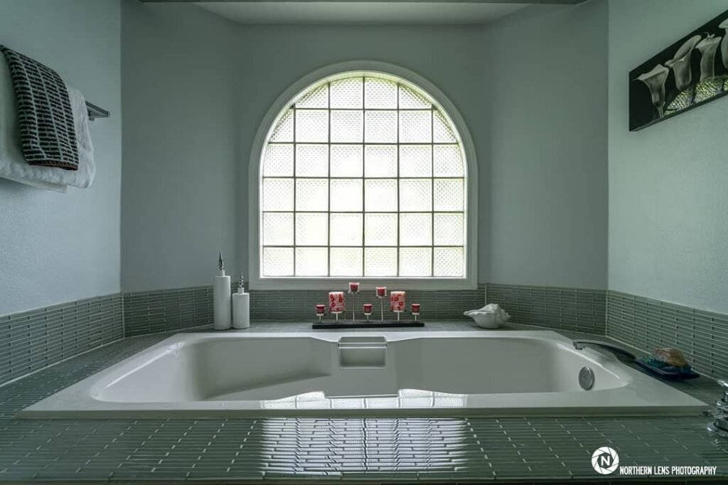 Serene bathroom featuring a large soaking tub bathed in natural light from an arched, grid-patterned window. Includes subtle decor and tiled accents.