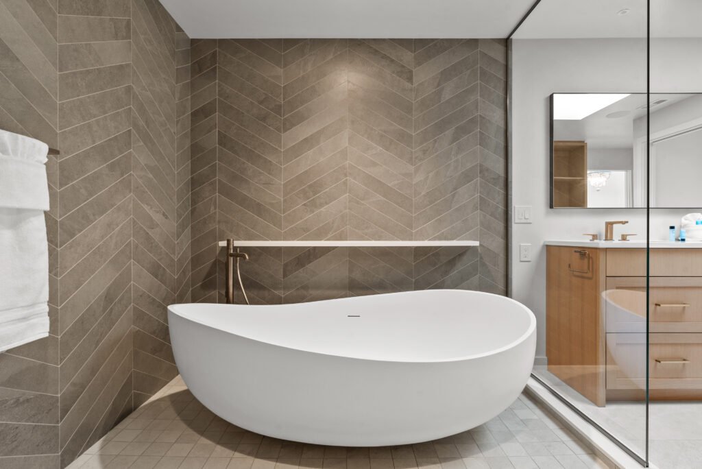 A beautifully designed modern minimalist bathroom featuring a sleek freestanding tub. The space is illuminated by soft, ambient lighting, highlighting the clean lines and elegant finishes. The neutral color palette and subtle textures contribute to a tranquil, spa-like atmosphere, perfect for relaxation.