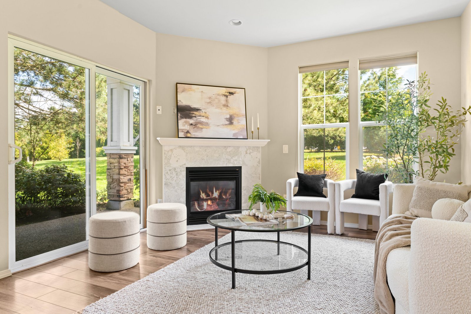 Staged living room with a fireplace, large windows, and a sliding glass door leading outside. The room contains a sofa, two armchairs, a glass coffee table, two ottomans, and artwork above the fireplace.
