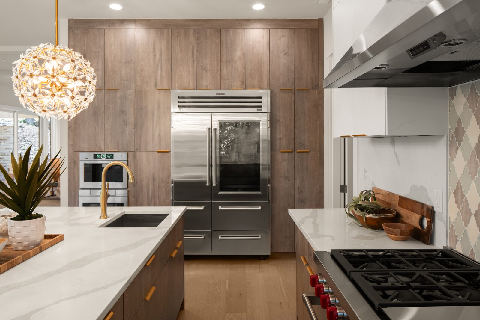 Modern luxury kitchen featuring light wood, floor-to-ceiling cabinets, a large stainless steel Sub-Zero refrigerator, a built-in oven, and a gas cooktop. A central island with a white quartz countertop and gold faucet is visible, with a decorative spherical crystal chandelier hanging above. The backsplash has a geometric tile pattern.