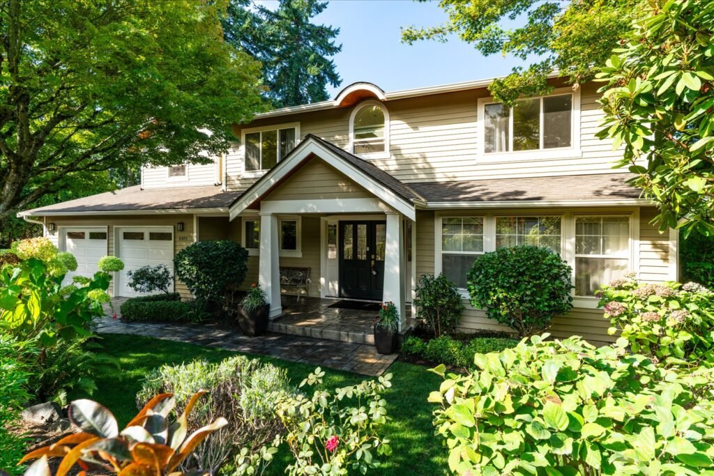 A detailed photo of a traditional two-story suburban home exterior, featuring light beige siding and a prominent covered entryway with a dark door. The property is framed by a lush, mature garden with a vibrant green lawn and large, deep-green trees, capturing the home's appealing curb appeal on a bright, sunny day.