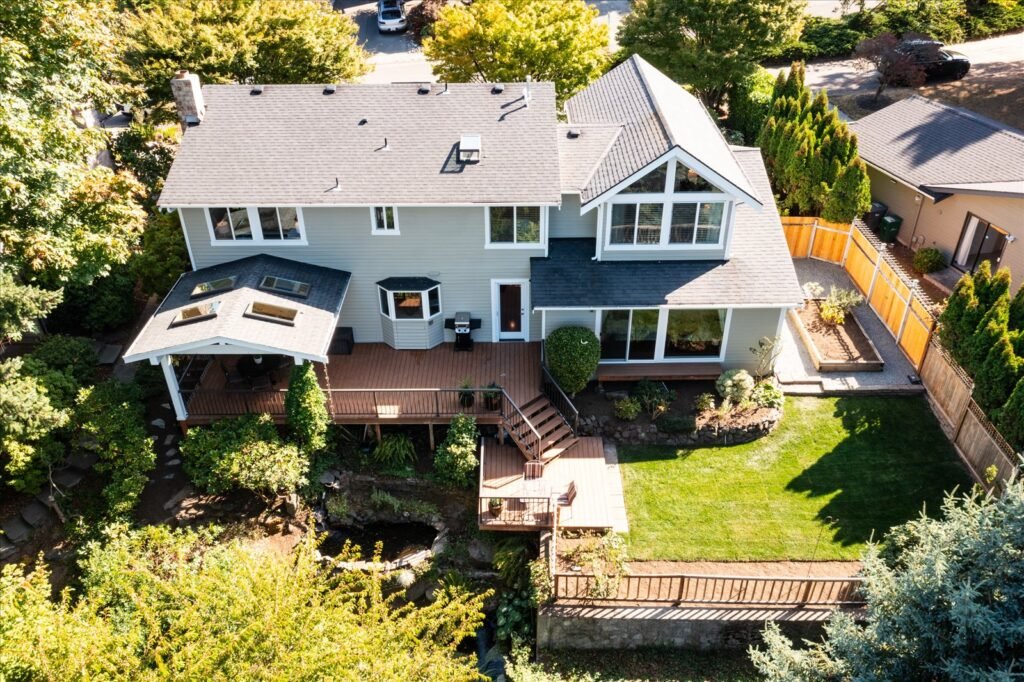 A clear, high-angle aerial photograph of the rear of a two-story suburban home with light gray siding and a dark gray roof. The house features a large, multi-level wooden deck, a covered patio area with skylights, and a spacious, well-maintained lawn. The backyard is surrounded by mature trees, highlighting the privacy and overall lot size from an elevated perspective.