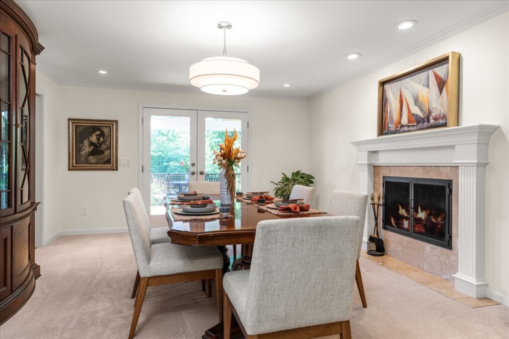A formal yet bright dining room with light beige walls and carpet, centered on a dark wood table set for four with light gray upholstered chairs. The room features a modern fireplace with a white mantel and a large, drum-style chandelier. White French doors at the far end open to an outdoor patio, enhancing the room's spacious and elegant feel.