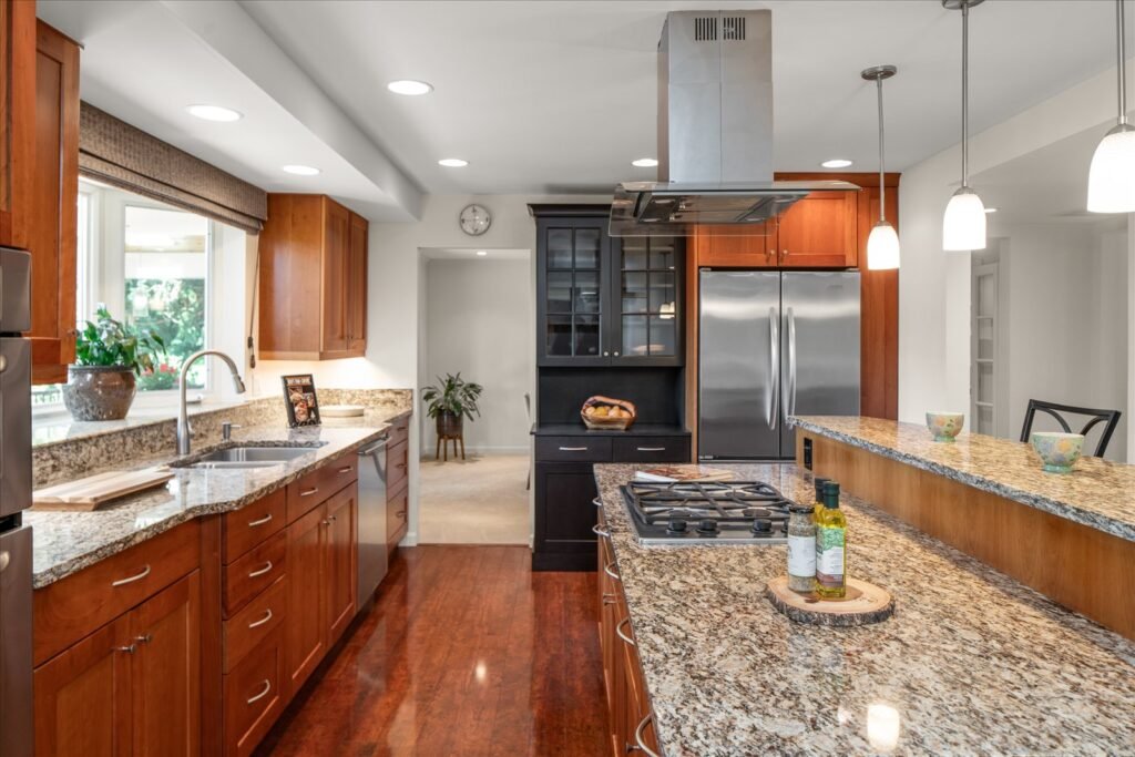 A brightly lit, galley-style kitchen featuring warm, cherry-toned wood cabinetry and multi-toned granite countertops. The space includes a stainless steel gas range, a matching hood vent, and a breakfast bar with modern pendant lighting. The rich hardwood floors contrast with the light walls, creating an inviting, professional-grade cooking space.