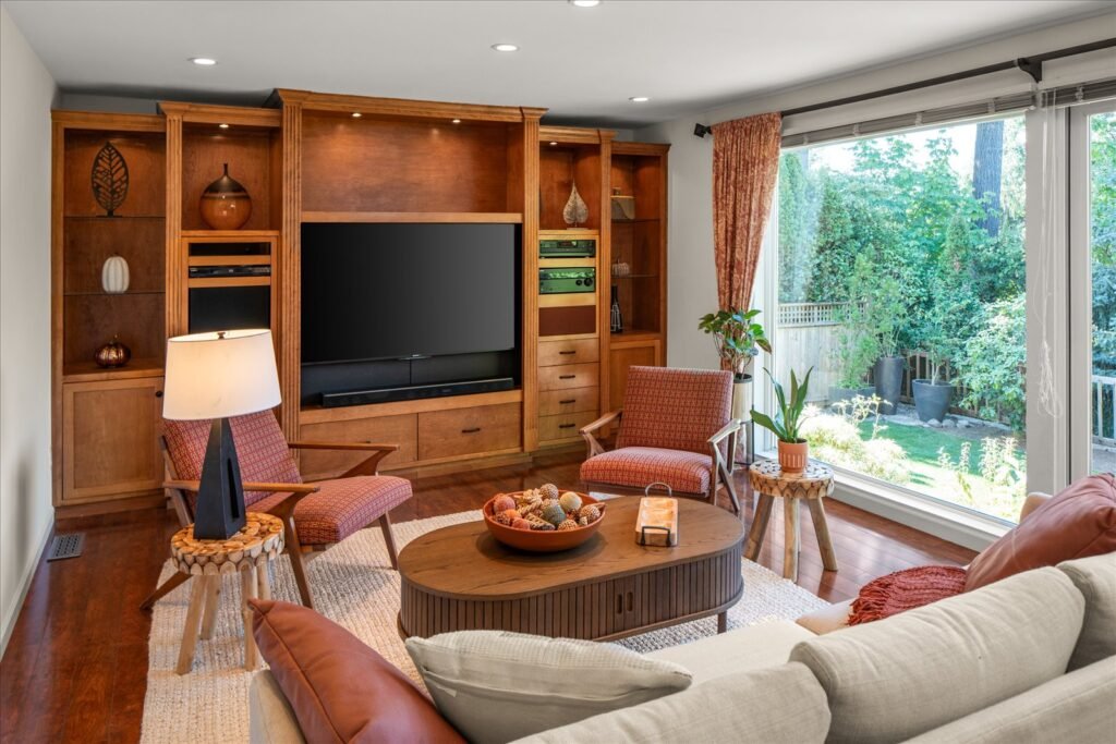 A bright, inviting family room with a light cream sofa in the foreground, facing custom-built, warm wood entertainment cabinetry and shelving that fills the back wall. The room is centered on a patterned area rug and features mid-century modern accent chairs, with a large picture window on the right offering a clear, expansive view of the lush, private backyard and patio.