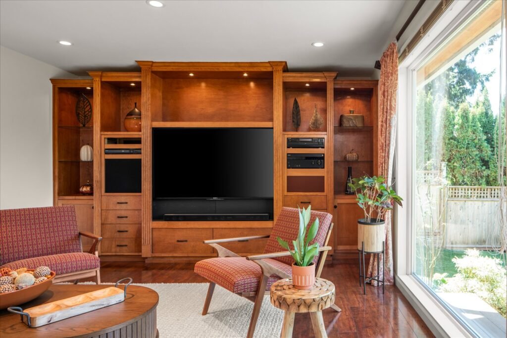 A cozy family room featuring a wall of rich, custom cherry wood built-in cabinetry with integrated lighting and open shelving surrounding a large, mounted television. Two mid-century modern style armchairs with patterned orange upholstery sit on dark hardwood floors next to a large window that provides a bright view of the private, fenced backyard and lawn.