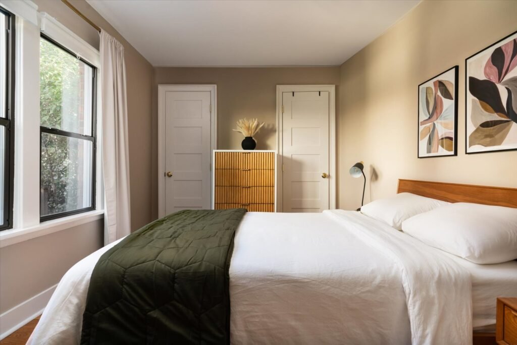 A cozy, stylish primary bedroom with warm greige-painted walls and dark wood flooring. The bed is dressed with crisp white linens and a dark olive green quilted throw. A window with black trim on the left provides ample natural light, and abstract art hangs over a mid-century modern wooden headboard, creating a clean, inviting, and peaceful atmosphere.
