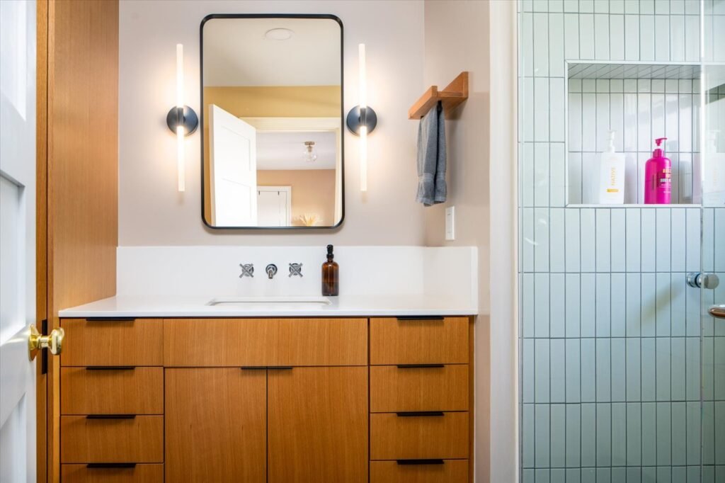 A modern and clean primary bathroom featuring a custom, mid-century style wood vanity with a long white solid-surface countertop and sleek black hardware. The wall-mounted faucets and illuminated rectangular mirror are flanked by vertical sconces. To the right, a glass-enclosed shower is visible, tiled in narrow, light blue-green vertical subway tiles with a recessed shelf.