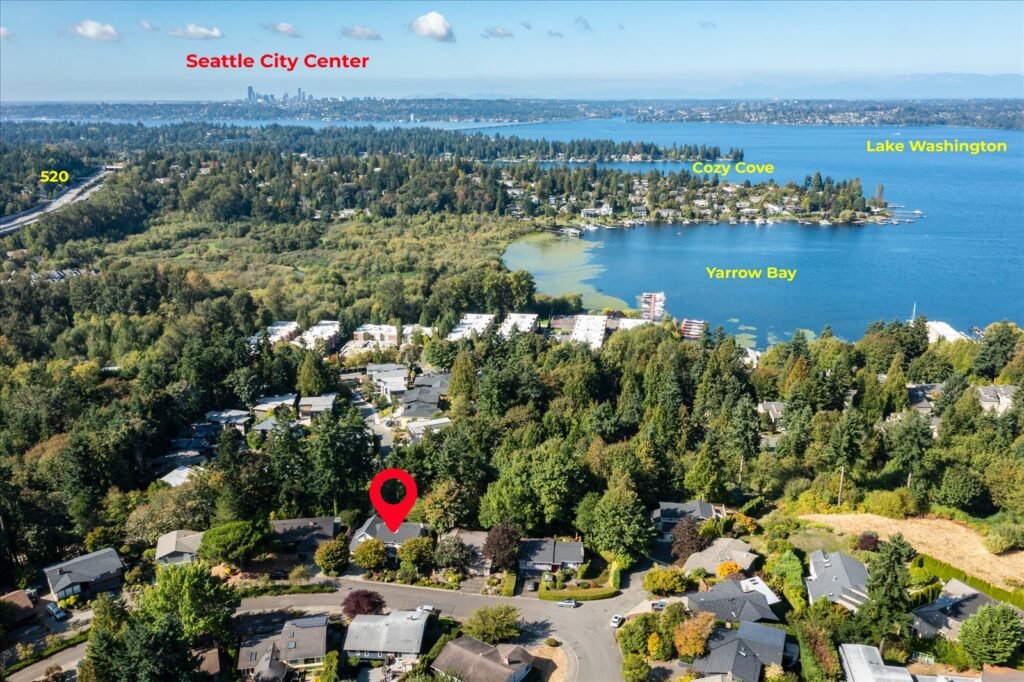 A striking, annotated aerial map view showing the location of a suburban home, marked with a red pin, situated in a lush, treed neighborhood bordering Lake Washington. In the background, the Seattle City Center skyline is clearly visible across the water, with the SR 520 bridge connecting the two shores. Key areas like Cozy Cove and Yarrow Bay are labeled, emphasizing the home's proximity to water and major metropolitan areas.