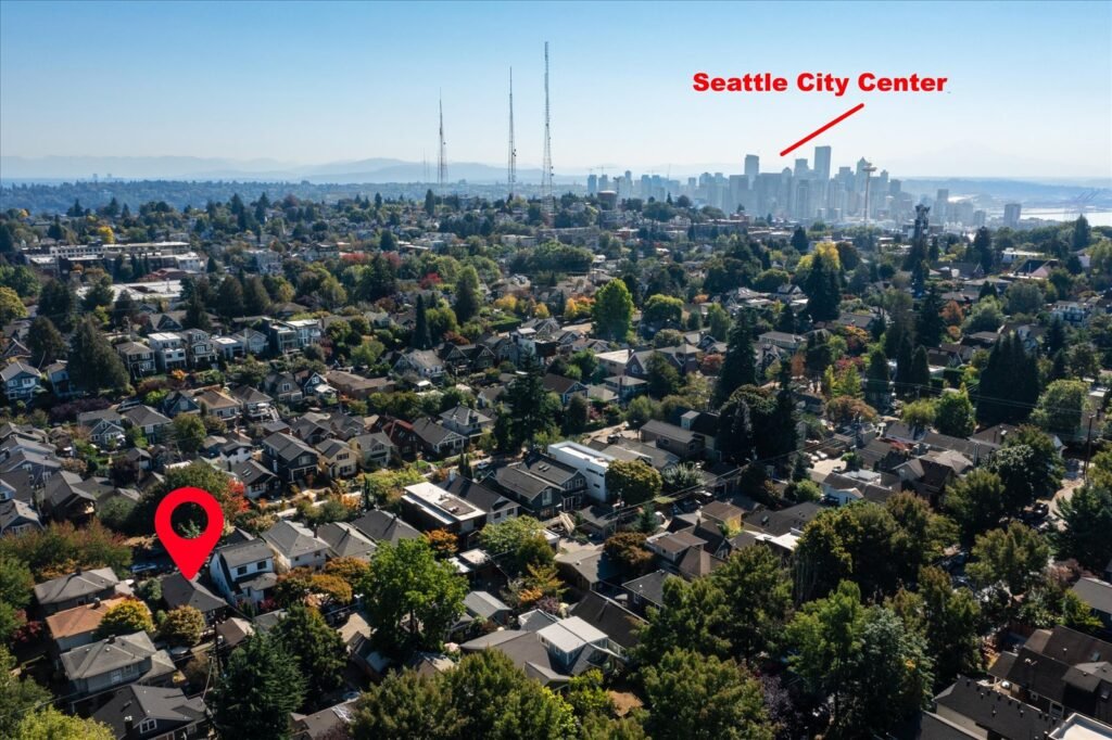 Aerial drone view looking southeast over residential Queen Anne neighborhood with tree-lined streets toward downtown Seattle skyline and Space Needle, mountains visible in background