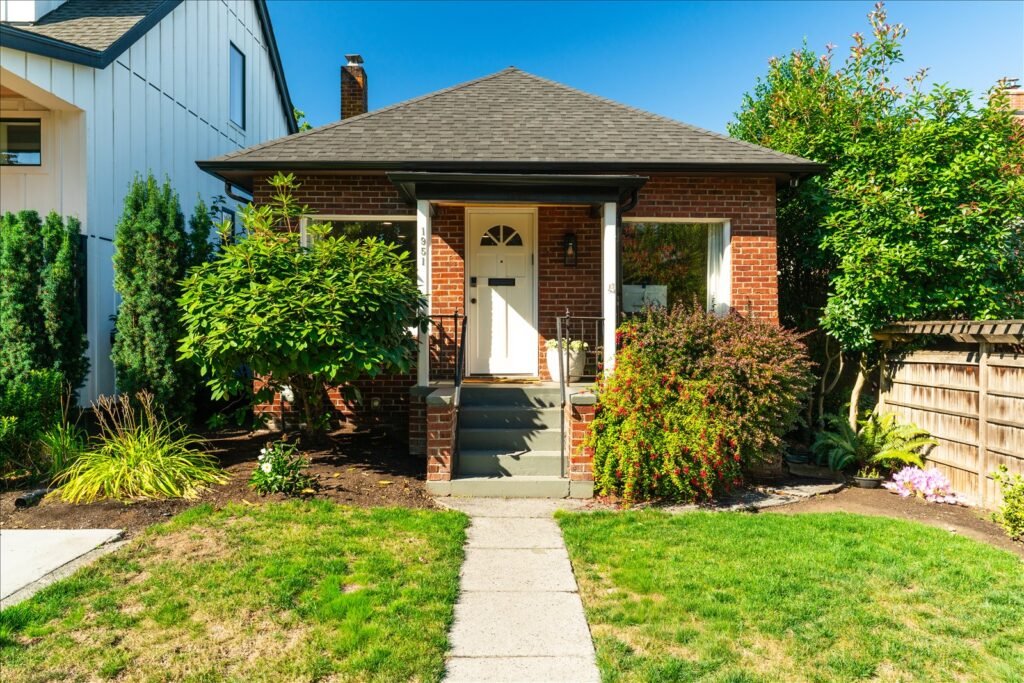 Front view of red brick single-story bungalow with white arched front door, concrete steps, mature landscaping, and manicured lawn under blue sky in Seattle neighborhood.
