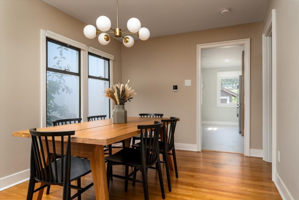 Modern dining room featuring live-edge wood table with six black Windsor chairs, brass sputnik chandelier, hardwood floors, and large windows with natural light on neutral walls