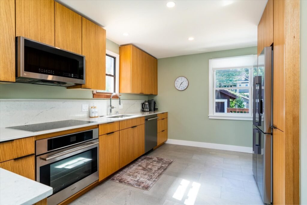L-shaped modern kitchen with floor-to-ceiling oak cabinets, white and gray quartz countertops, stainless steel appliances including wall oven and microwave, large window, and sage green accent wall