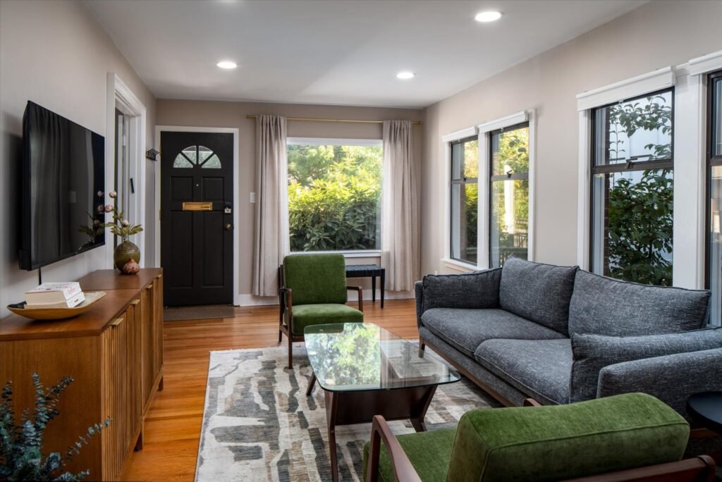 Bright living room with hardwood floors, gray upholstered sofa, green velvet accent chairs, glass coffee table, oak credenza, wall-mounted TV, black front door with arched window detail, and large windows with white trim