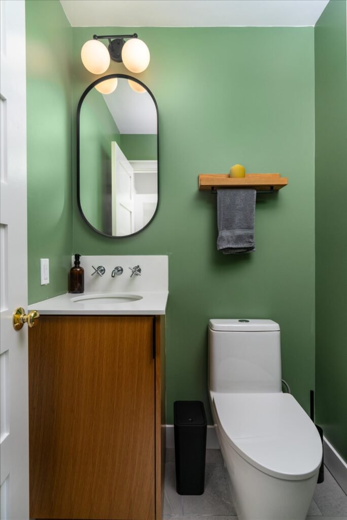 compact powder room with sage green walls, oval black-framed mirror, oak wood vanity with white countertop, wall-mount chrome faucets, and brass door hardware