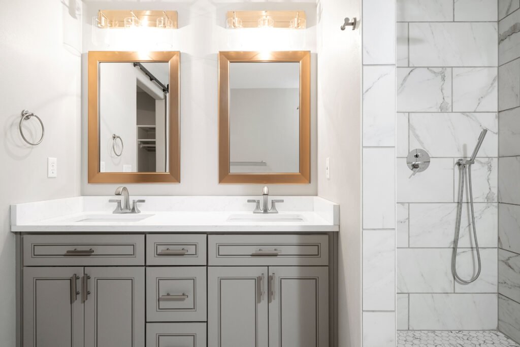 A bright, modern master bathroom featuring a double vanity with grey shaker cabinets and a white quartz countertop. A large walk-in shower with white marble-look tiles is visible on the right, contrasted by brushed bronze mirrors and gold light fixtures, emphasizing a clean and luxurious design.