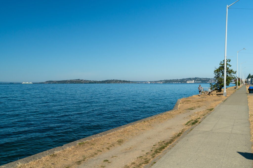 Jogger on Alki Beach waterfront walking path with Puget Sound and Olympic Peninsula views.