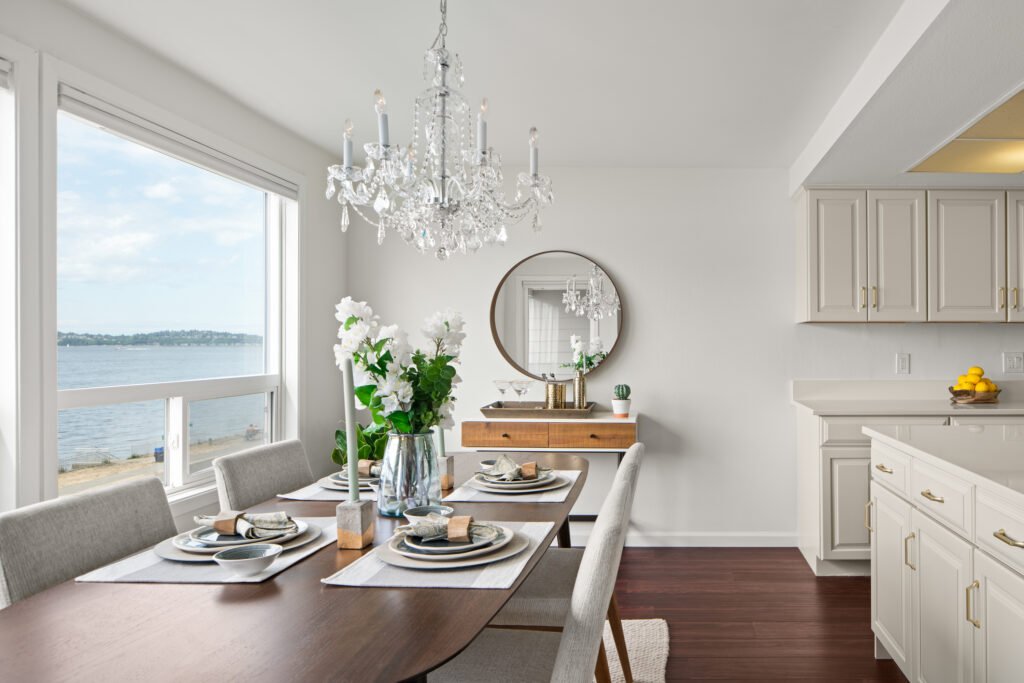 Elegant waterfront condo dining room with crystal chandelier, mid-century modern sideboard, large round mirror, and Puget Sound water views through windows