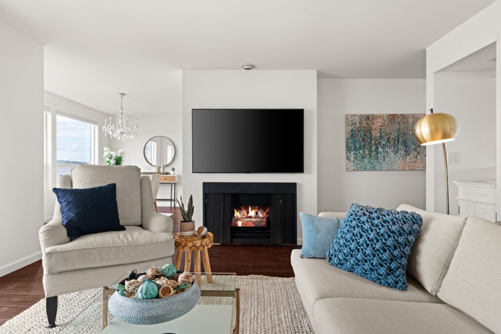 Modern Alki Beach condo living room with black fireplace, beige furniture, coastal blue accent pillows, and crystal chandelier with water views through windows
