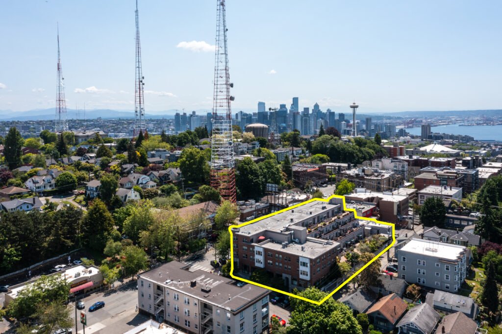Aerial drone view of Seattle apartment building with yellow outline showing property location, Space Needle and downtown Seattle skyline visible in background, surrounded by residential neighborhoods and trees
