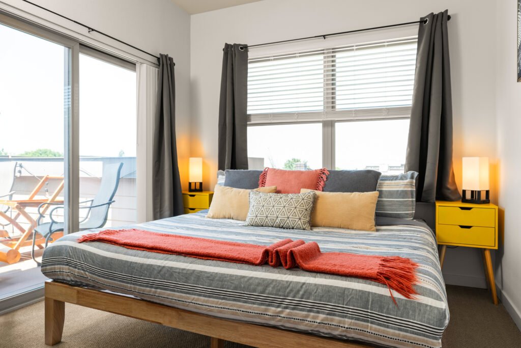 Modern bedroom with platform bed featuring striped gray bedding and coral accents, bright yellow nightstands with lamps, sliding glass door to balcony, and hardwood floors
