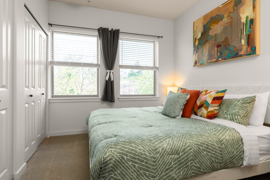 Bright bedroom with white tufted headboard, colorful geometric pillows, sage green bedding, two windows with blinds, hardwood-style flooring, and modern artwork