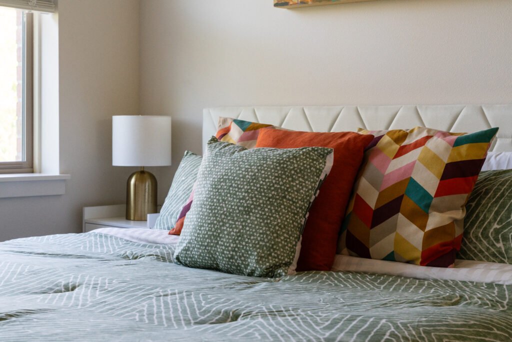Close-up of bed with white tufted headboard, layered pillows in sage green, coral, and geometric patterns, sage green bedspread with textured weave
