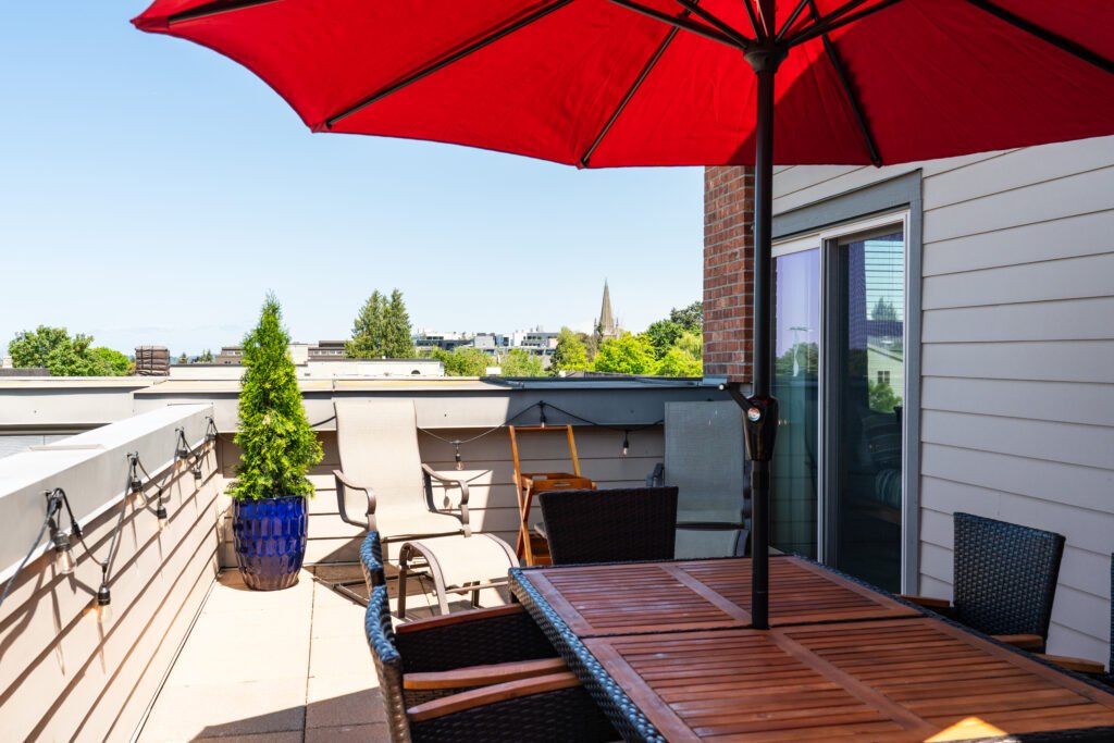 Bright rooftop deck with red umbrella, wooden lounge chairs, patio furniture, city skyline views, and potted plants under clear blue sky in Seattle