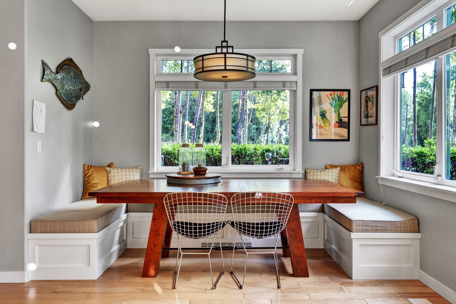 Modern dining nook with built-in white bench seating with cushions, dark wood table, wire metal chairs, gray walls, large windows with garden views, hardwood floors, and drum pendant light fixture
