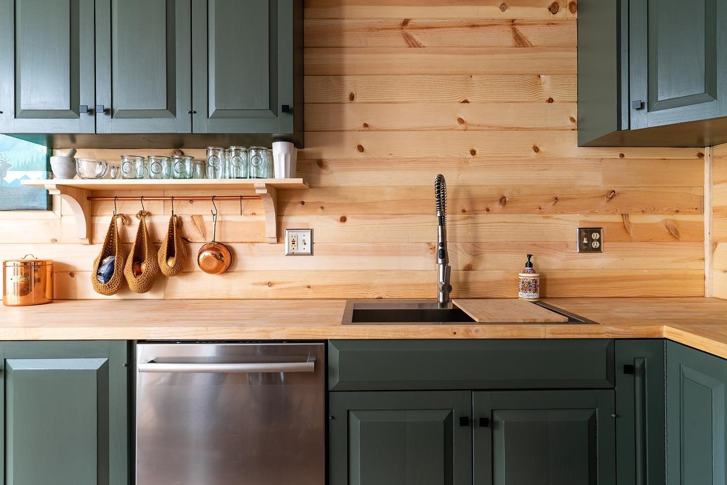 Modern kitchen with sage green shaker cabinets, natural pine wood plank backsplash and countertops, undermount black sink with modern faucet, open shelving with glassware, woven basket storage hooks, stainless steel dishwasher
