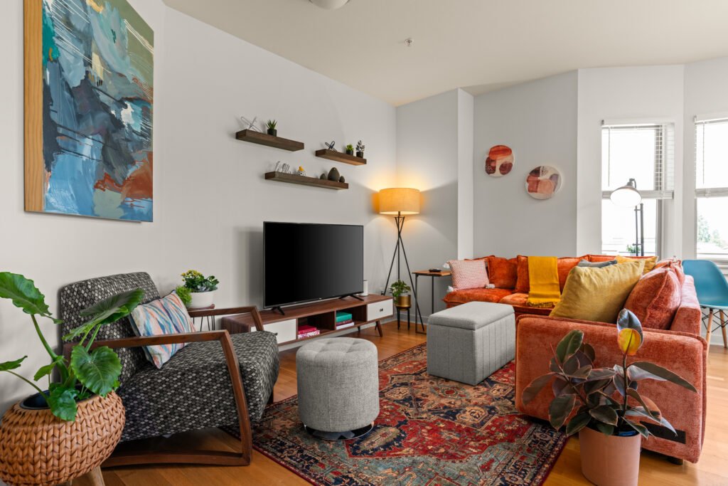 Colorful living room with burnt orange velvet sofa, patterned rug, gray upholstered chair, wall-mounted TV on wood console, floating shelves, modern artwork, and potted plants